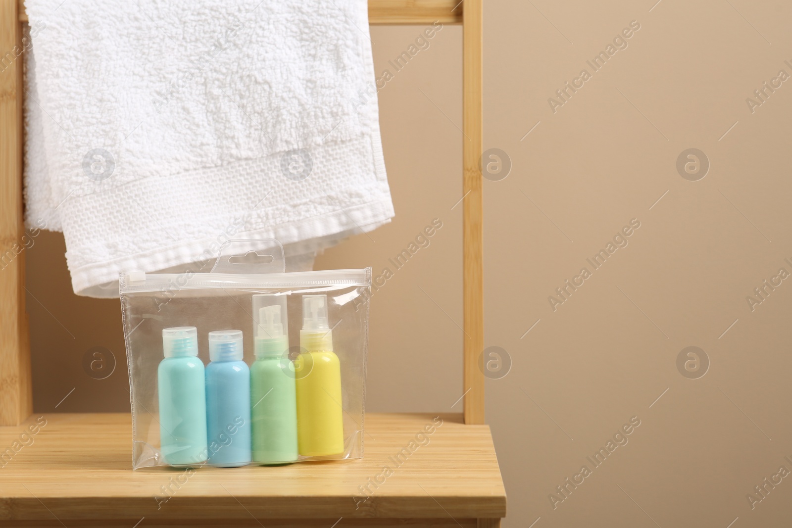 Photo of Cosmetic travel kit in plastic bag and towel on wooden chair against beige wall, space for text. Bath accessories