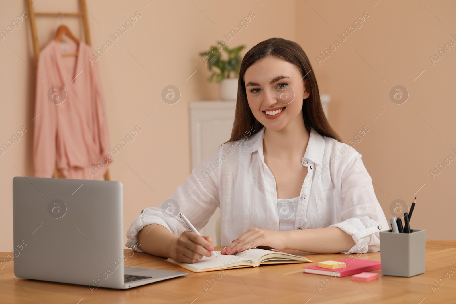 Photo of Happy young woman sitting near notebook at wooden table indoors