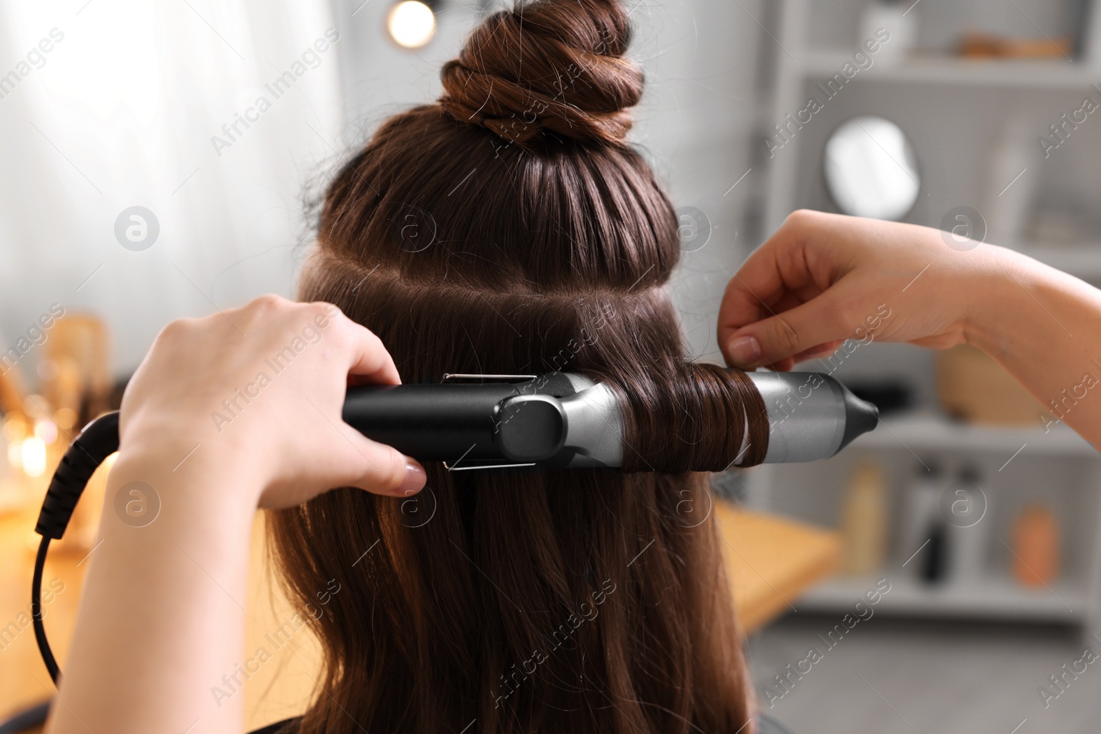 Photo of Hairdresser using curling hair iron while working with woman in salon, closeup