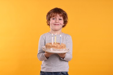 Photo of Birthday celebration. Cute little boy holding tasty cake with burning candles on orange background