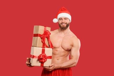 Photo of Muscular young man in Santa hat with Christmas gift boxes on red background