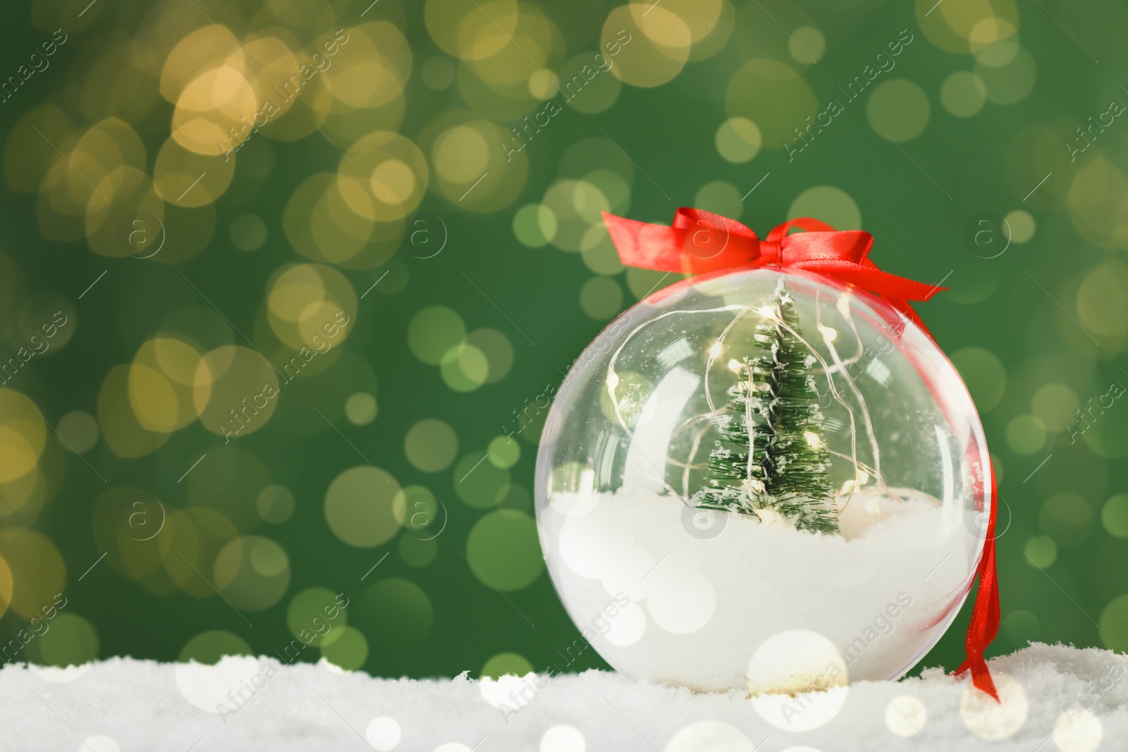 Image of Beautiful transparent Christmas ornament with small fir tree and fairy lights on snow against green background, bokeh effect. Space for text