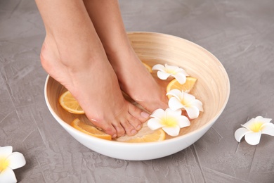 Photo of Woman soaking her feet in bowl with water, orange slices and flowers on grey background, closeup. Spa treatment