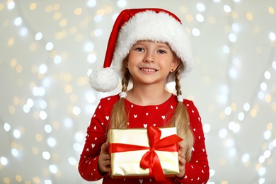 Photo of Happy little child in Santa hat with gift box against blurred festive lights. Christmas celebration