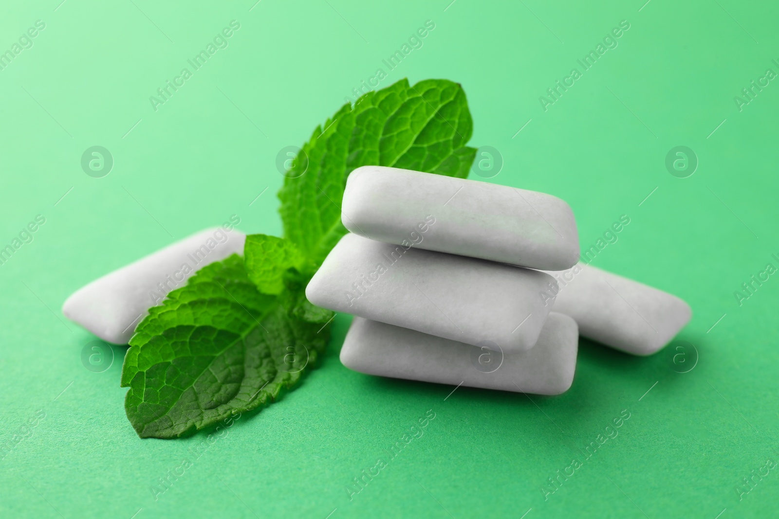 Photo of Tasty white chewing gums and mint leaves on green background, closeup