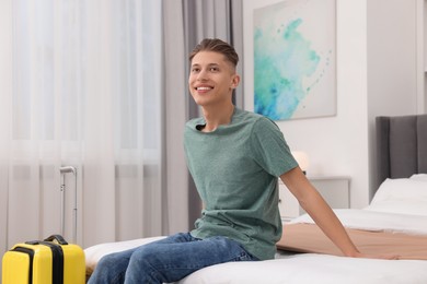 Photo of Smiling guest with suitcase relaxing on bed in stylish hotel room