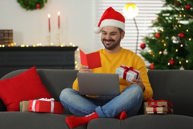 Photo of Celebrating Christmas online with exchanged by mail presents. Man in Santa hat with greeting card and gift box during video call on laptop at home
