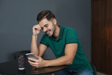 Photo of Young man with cup of coffee and smartphone laughing at table indoors