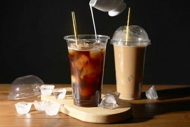 Pouring milk into takeaway cup with refreshing iced coffee at wooden table against black background