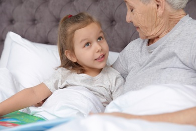 Cute girl and her grandmother reading book on bed at home