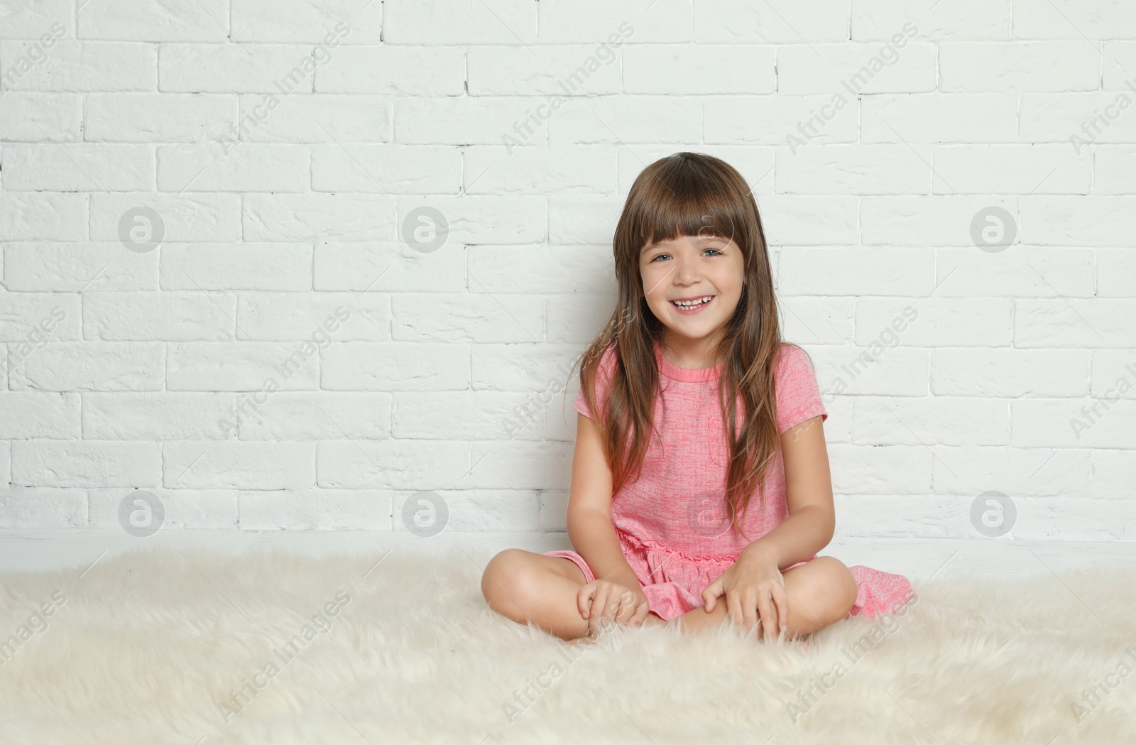 Photo of Cute little girl sitting on fur rug against brick wall. Space for text