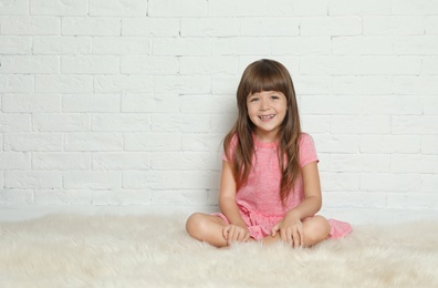 Cute little girl sitting on fur rug against brick wall. Space for text