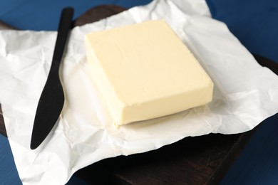 Photo of Block of tasty butter in open foil packaging and knife on blue wooden table, closeup