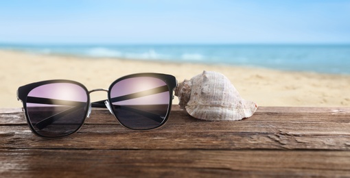Shell and stylish sunglasses on wooden table near sea with sandy beach. Banner design