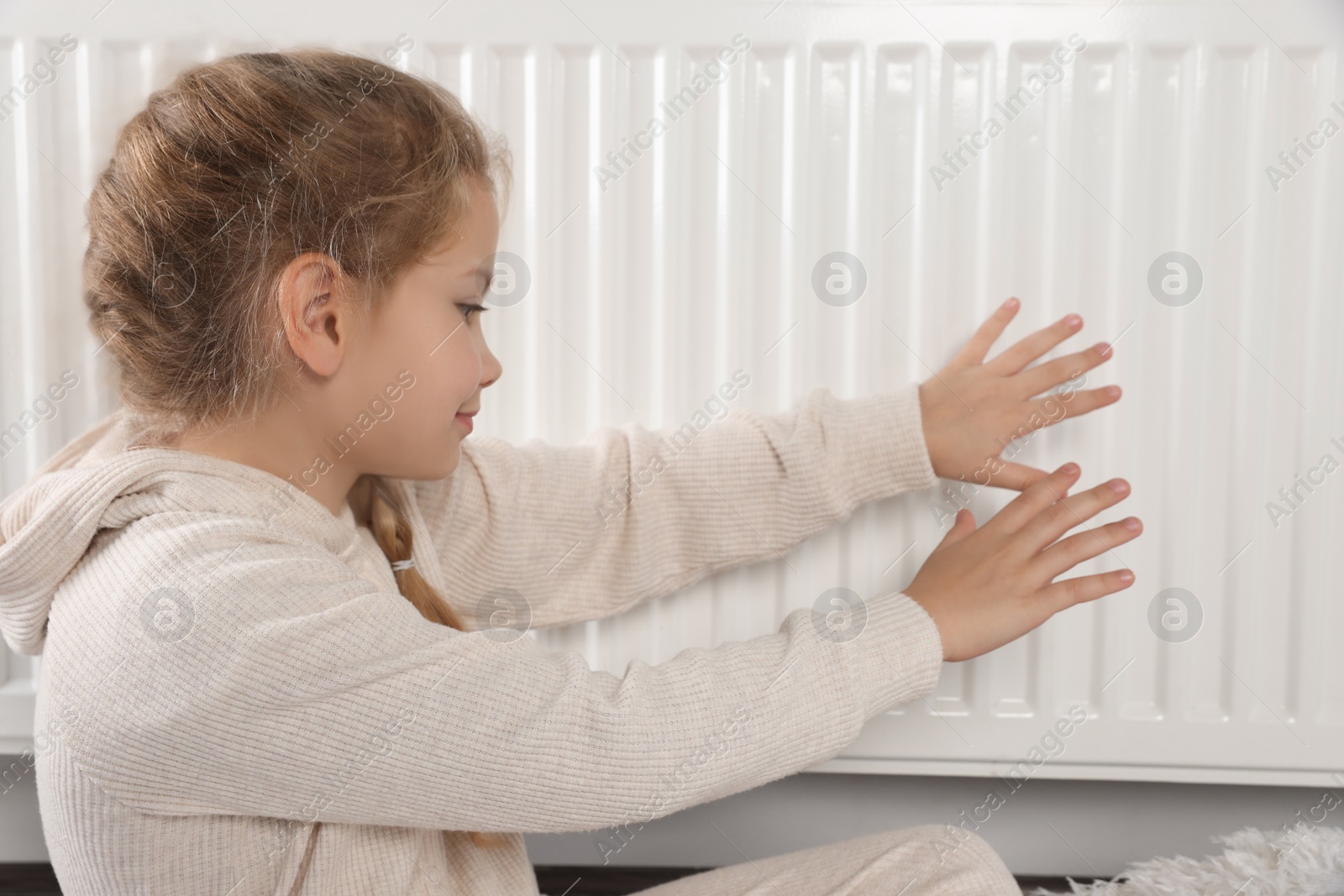 Photo of Little girl warming hands near heating radiator indoors