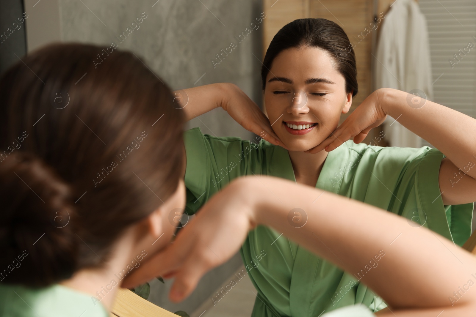 Photo of Young woman massaging her face near mirror in bathroom