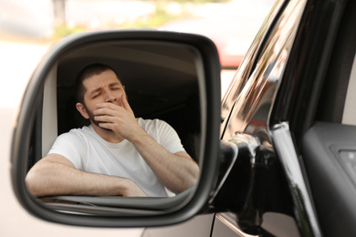 Photo of Tired man yawning in his auto, view through car side mirror