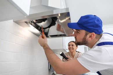 Photo of Worker repairing modern cooker hood and woman holding flashlight in kitchen