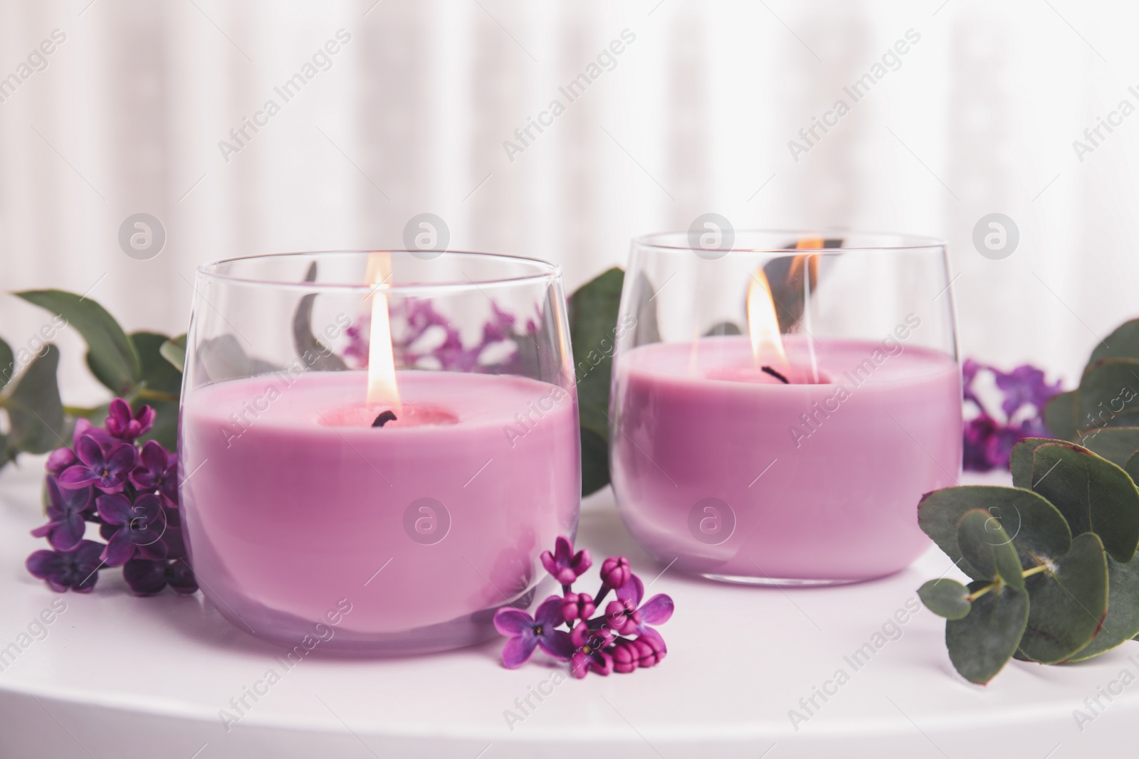 Image of Burning candles in glass holders and flowers with leaves on white table