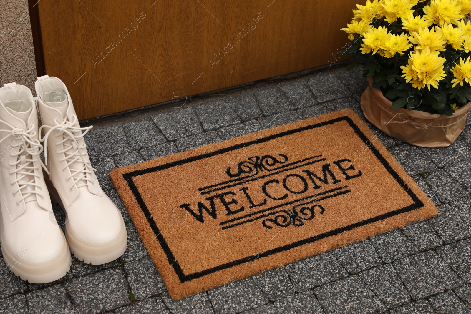 Photo of Doormat with word Welcome, stylish boots and beautiful flowers on paving stone near entrance