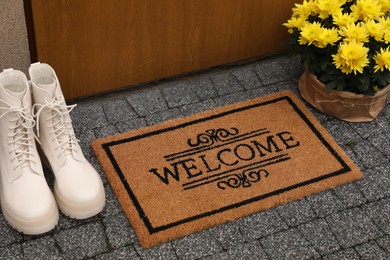 Photo of Doormat with word Welcome, stylish boots and beautiful flowers on paving stone near entrance