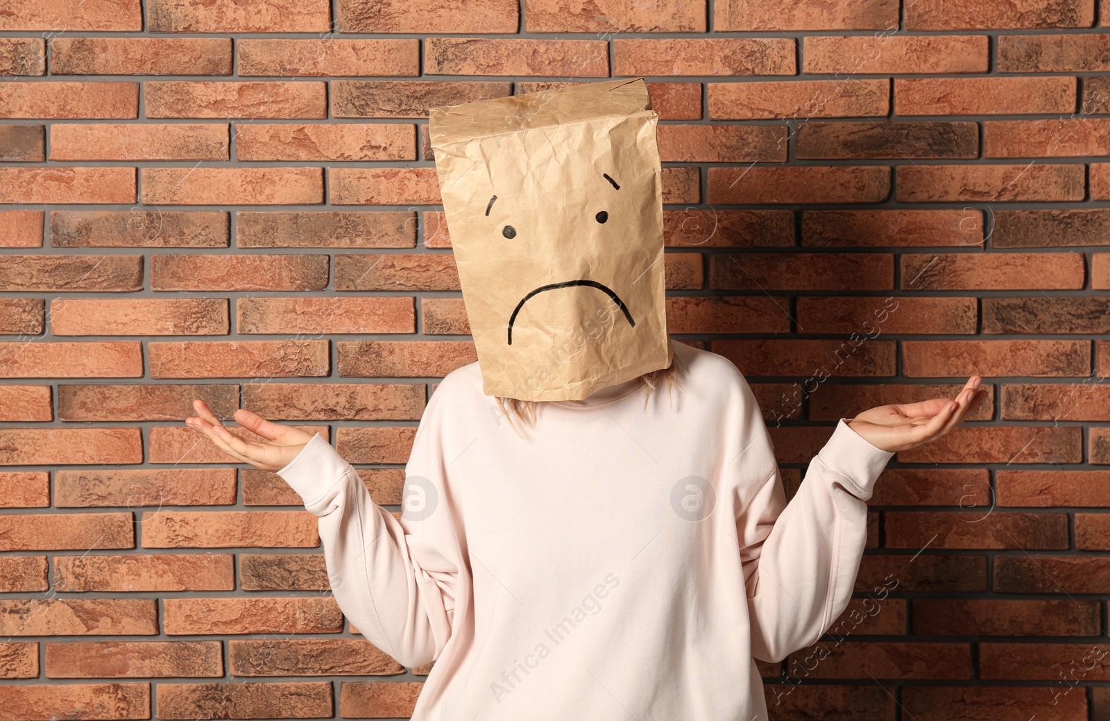 Photo of Woman wearing paper bag with drawn sad face near brick wall