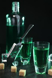 Photo of Absinthe in shot glasses, spoon, brown sugar and ice cubes on gray table against dark background, closeup. Alcoholic drink