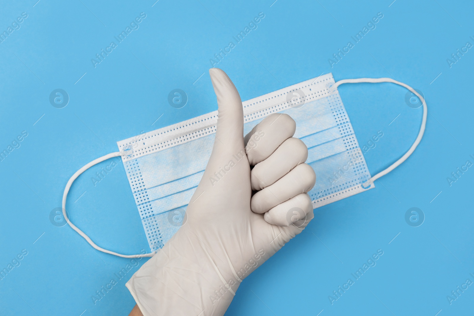 Photo of Doctor in medical gloves showing thumb up gesture near protective mask on light blue background, top view
