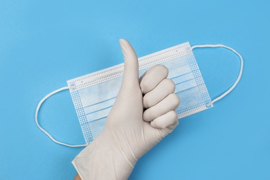 Doctor in medical gloves showing thumb up gesture near protective mask on light blue background, top view