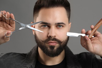 Handsome young man with mustache holding blade and scissors on grey background