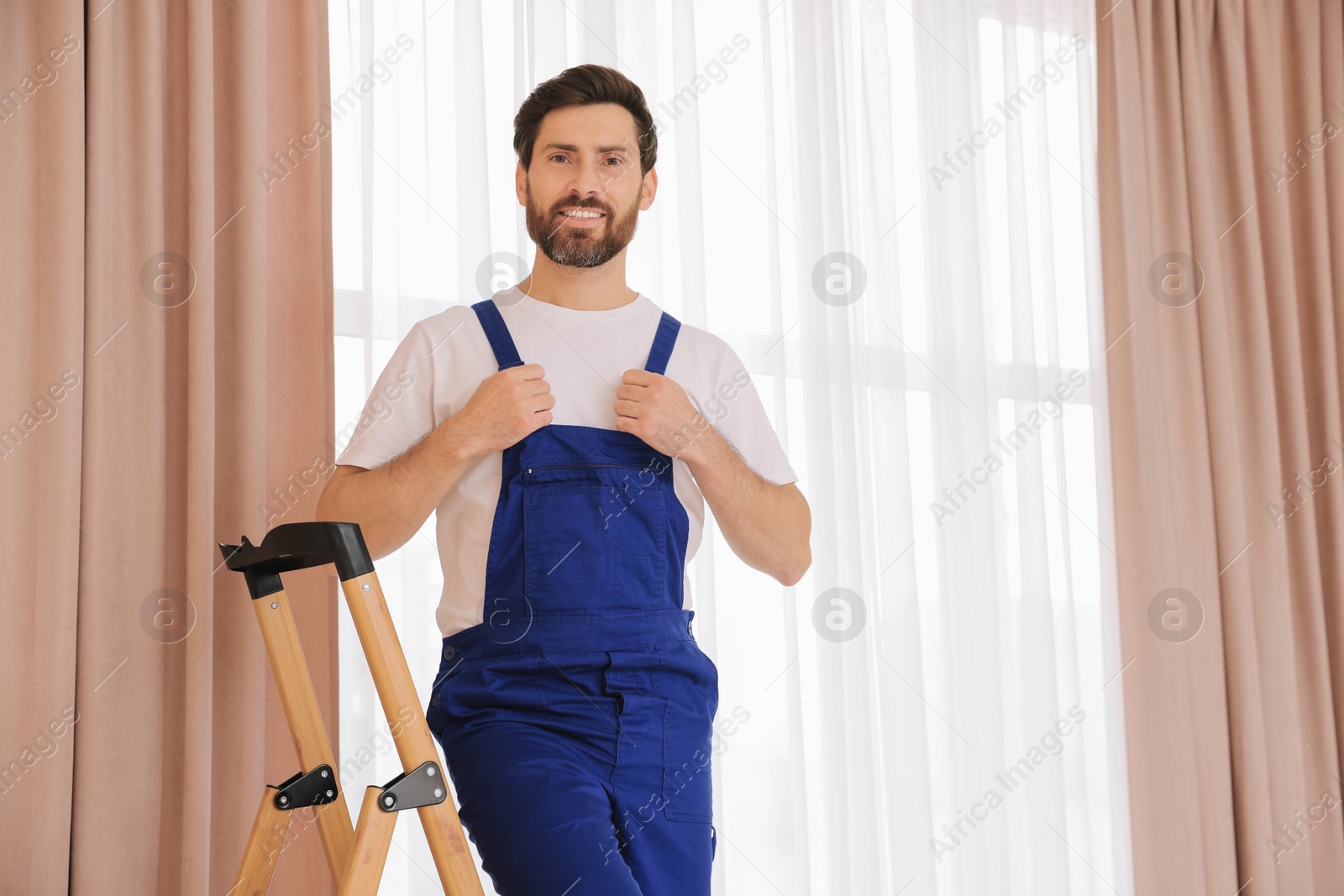 Photo of Worker in uniform standing on wooden folding ladder near window curtains indoors