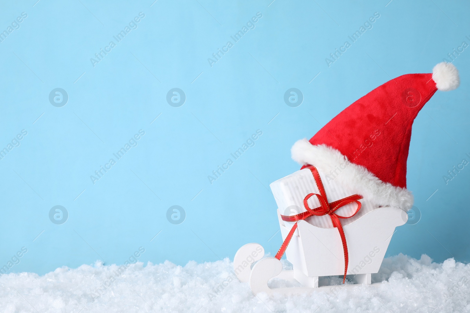 Photo of White wooden sleigh with Christmas gift box, Santa hat and artificial snow on light blue background, space for text