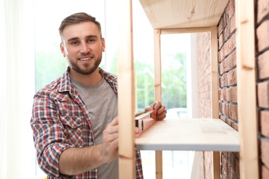 Young man working with construction level near wooden shelving unit indoors