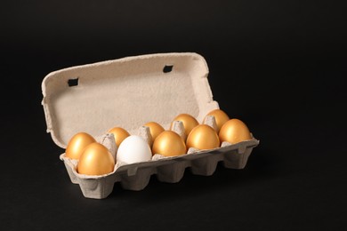 Photo of Ordinary chicken egg among golden ones in box on black background