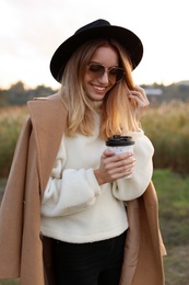 Beautiful young woman with cup of coffee wearing stylish autumn clothes outdoors