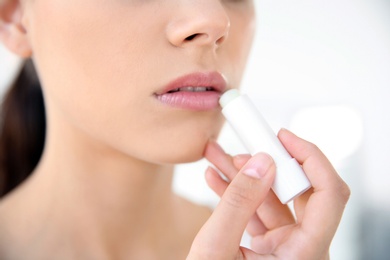 Young woman applying balm on her lips against light background, closeup