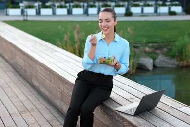 Smiling businesswoman eating from lunch box near laptop outdoors. Space for text