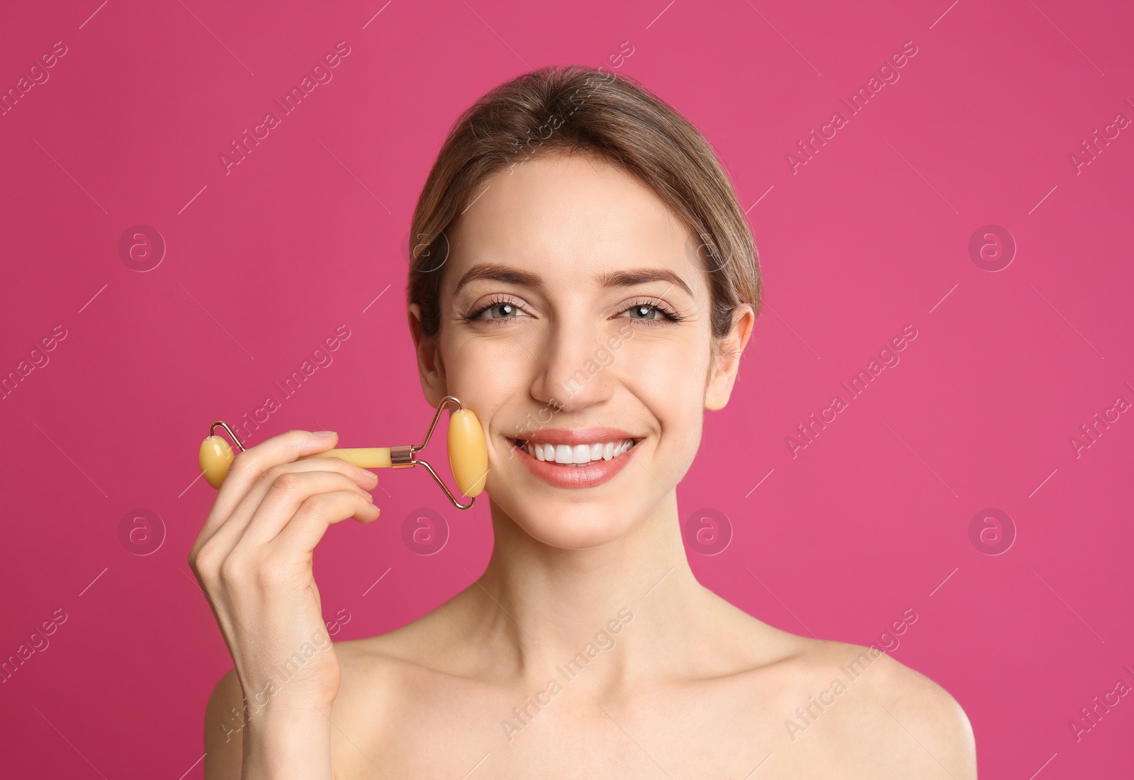 Photo of Young woman using natural jade face roller on pink background