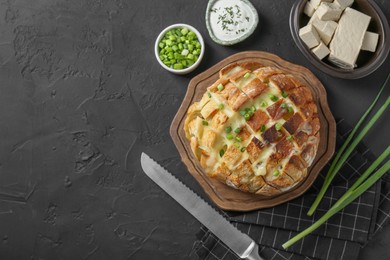 Photo of Freshly baked bread with tofu cheese, green onions, sauce and knife on black table, flat lay. Space for text