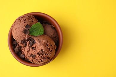 Bowl of tasty ice cream with chocolate chunks and mint on yellow background, top view. Space for text