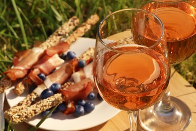 Photo of Wooden board with glasses of delicious rose wine and food on green grass outdoors, closeup