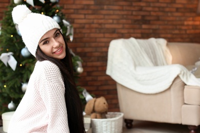 Beautiful young woman in hat near Christmas tree at home