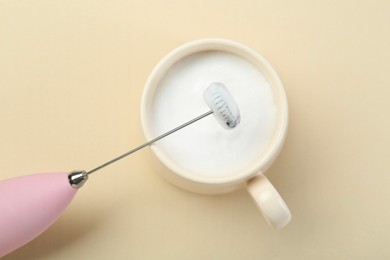 Photo of Mini mixer (milk frother) and cup of whipped milk on beige background, top view