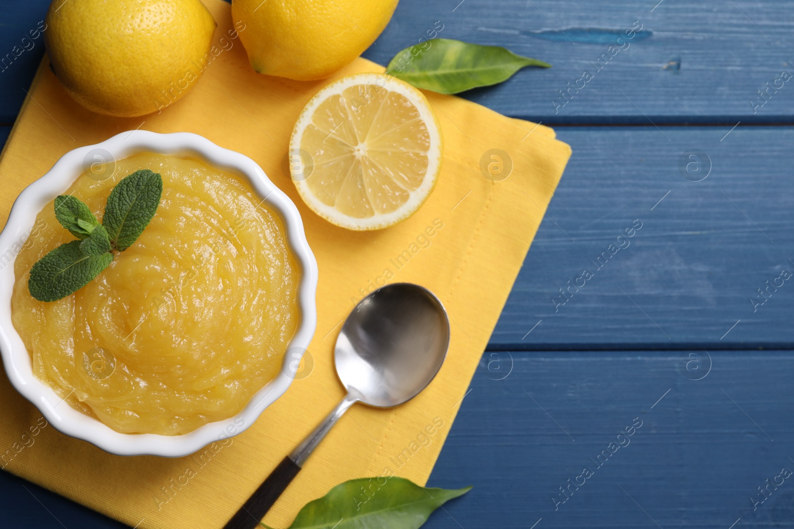 Photo of Delicious lemon curd in bowl, fresh citrus fruits and spoon on blue wooden table, flat lay. Space for text