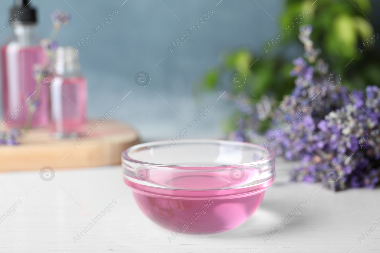 Photo of Bowl of lavender essential oil on white wooden table against blue background. Space for text