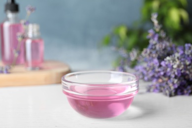 Photo of Bowl of lavender essential oil on white wooden table against blue background. Space for text