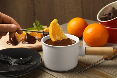 Photo of Woman dipping slice of orange into fondue pot with milk chocolate at wooden table, closeup