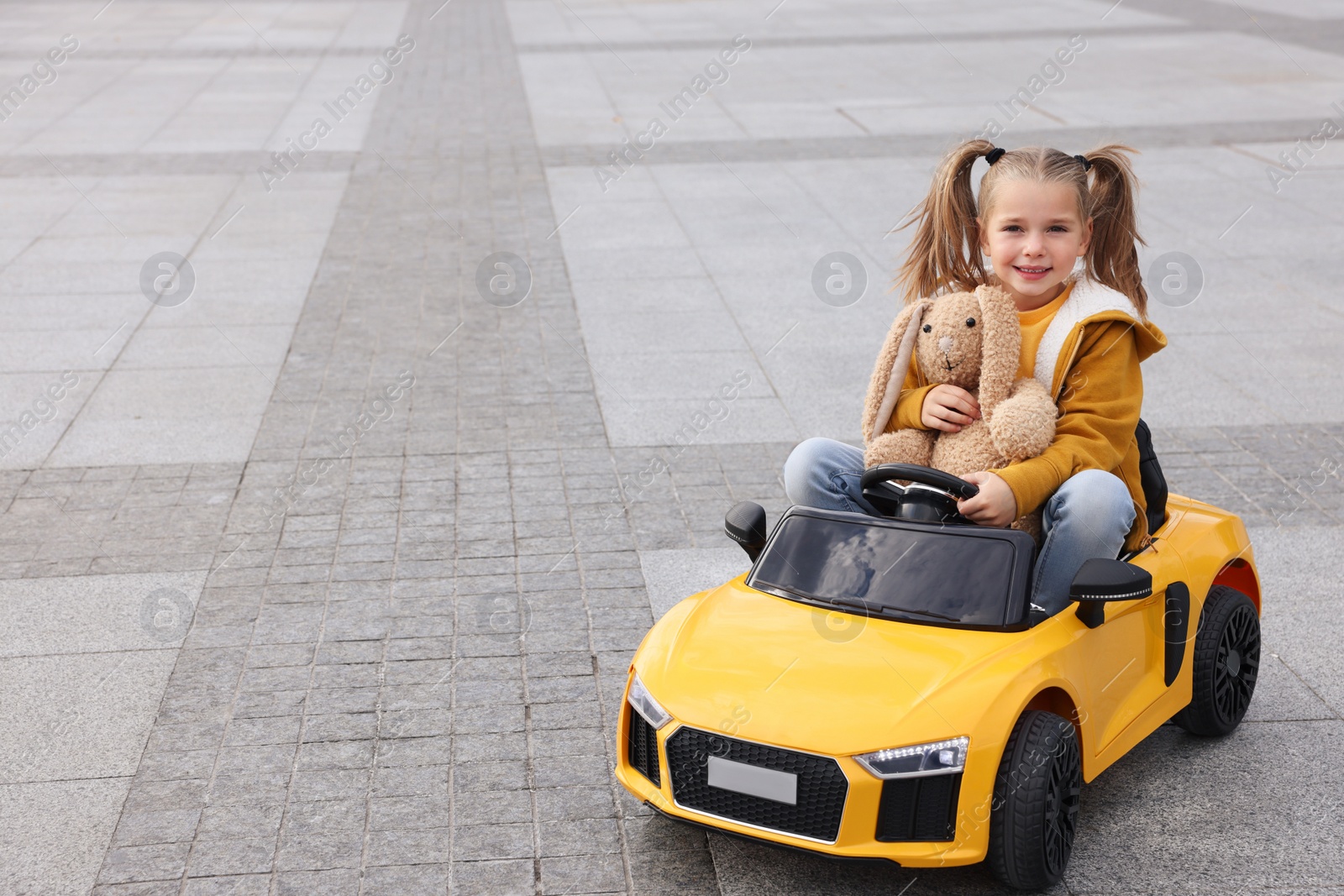 Photo of Cute little girl with toy bunny driving children's car on city street. Space for text