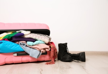 Suitcase with clothes and boots on floor against white wall, space for text. Winter vacation