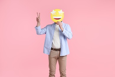 Photo of Man holding emoticon with stars instead of eyes and showing peace sign on pink background